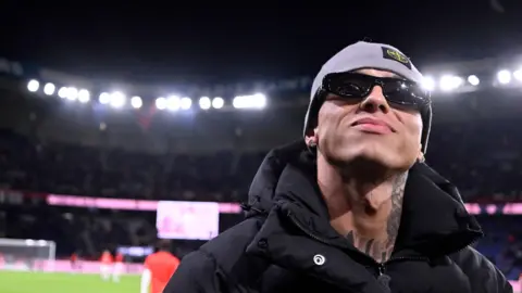 Getty Images Central Cee, wearing sunglasses, stands on the pitch of Parc Des Princes in France, as he attends a football match in September 2024