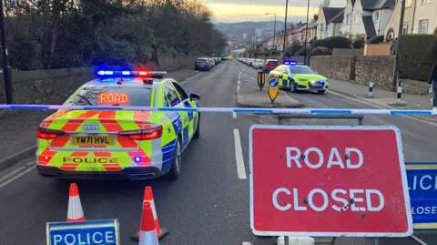 Police cordon on a street, with police vehicles and road closed signs in red and white