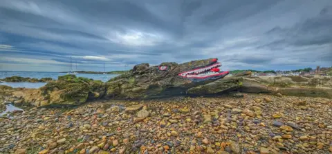 Mangela Coia The face of a crocodile painted onto a long rock on a pebble beach