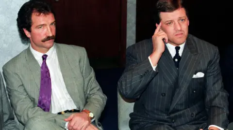 SNS Graeme Souness and David Murray at a press conference. Moustachioed Souness wears a patterned green suit, a purple tie and white shirt. Murray wears a pinstriped black suit, a white shirt and what appears to be a white and black polka dot tie, though his broad lapel covers most of it.
