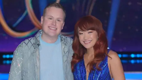 Josh Jones and his dance partner Tippy Packard stand smiling on the ice, both wearing sparkly blue outfits.