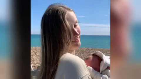 Morgan Betchley is a teenager with long blonde hair. She stands on a beach in the sunshine cradling her newborn son. 