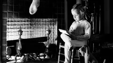 Getty Images A black and white image of a girl of about five or six years old, sitting on a chair beside a fireplace. She is resting her right leg across her left knee and has a notepad resting on the leg on which she is writing. Above the fireplace is hanging the bottom half of a stocking