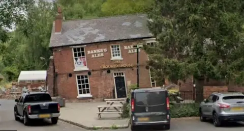 Google An image of redbrick building with slated roof leaning heavily to the left hand side. Signage on it reads Banks's Ales and The Crooked House. Trees are in front and behind with three cars parked outside