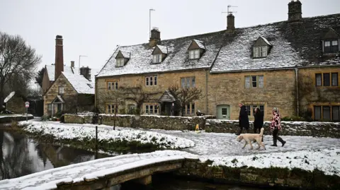 A wide image of the Cotswold village of Lower Slaughter after it had a covering of snow. In the foreground is a waterway with a stone bridge over it, while a row of traditional honeycomb stone houses can be seen. Someone is walking a cream-coloured dog in front of the houses