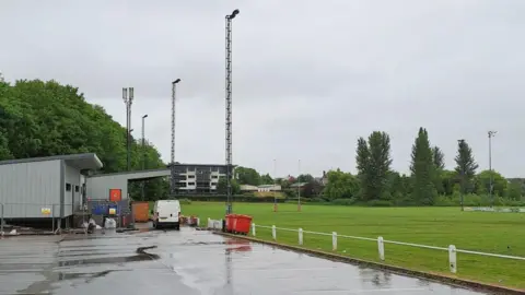 A rugby pitch with car park in the foreground. There is a block of flats in the distance, overlooking the club, and a club house and spectator stand to the left hand side of the image.