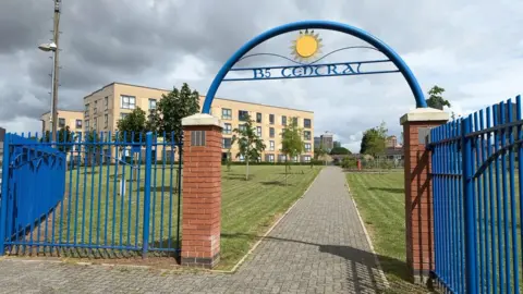 The entrance to B5 Central with blue metal railings either side of red brick pillars, with a blue metal arch over them and the letters B5 Central sculpted out of metal. A grey paved path leads to a beige brick building.