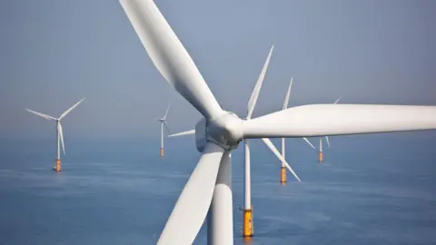 A close up of an offshore wind turbine hub and blades with other turbines in the background. The sea below is flat and calm.