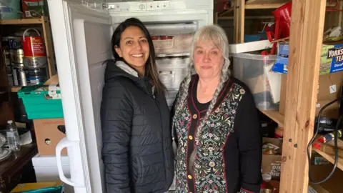 Two women standing side by side in a garage. They are positioned next to an upright freezer with the door open, revealing neatly stacked containers inside. The woman on the left is wearing a dark padded jacket and has long dark hair. The woman on the right is wearing a patterned cardigan with floral detailing and has long grey hair styled in braids. The surrounding area contains various storage items, including plastic bins, boxes, shelves, and household supplies.