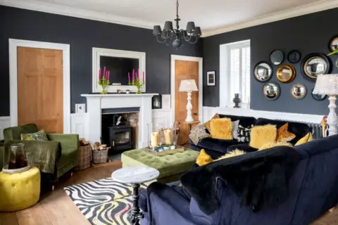 IWC Media Inside The Schoolhouse. The living room has black walls and dark coloured furniture, with a white fire place in the centre of the back wall. 