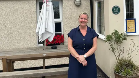 Lois Bradburn A pub landlady - a middle-aged woman with blonde hair who is wearing a button up navy dress with short sleeves - stands in front of a pub. There is a picnic bench with a parasol behind her as well as some plants.