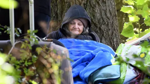 Alan Lewis Ann Hillman in a blue sleeping back sits against a tree sleeping. She is wearing a dark coat with the hood up, and a stripped wooly hat. 