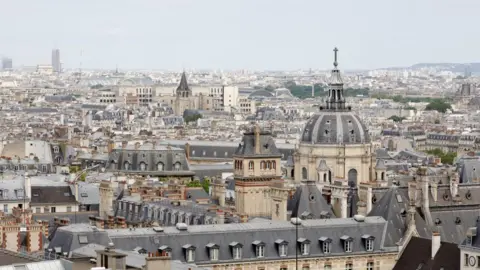 The World Monuments Fund A bird's eye view of Paris in France, the grey roof tops of various buildings can be seen including spire tops, flat roods and dome shaped roofs. Most of the buildings have light stone coloured walls.