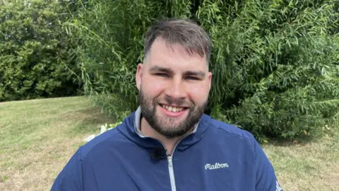 A man, Jonothan Crooks. He is pictured smiling outside in a garden. He is wearing a navy rain jacket; he has brunette hair and a beard.