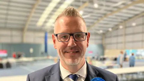 BBC John Campion with short, spikey grey hair. He is pictured in a suit in a sports hall. 