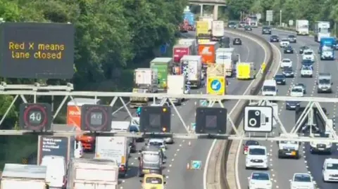 National Highways A traffic camera view of a congested motorway. Some of the lanes are coned off and an ambulance can be seen in the distance.