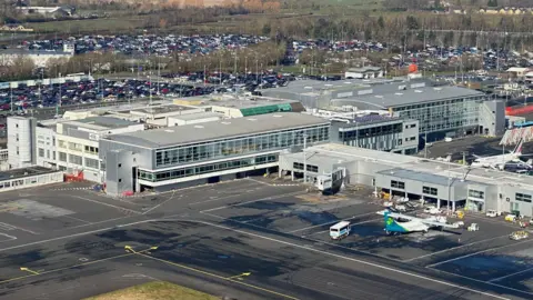 Aerial view of Newcastle Airport. A large plane can be seen on tarmac next to a large terminal building, which is boxy and flat 