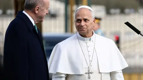 Reuters Pope Leo smiles as he looks towards Erdogan while wearing a white robe with a silver cross on a necklace while standing outside the Presidential Palace on Thursday.