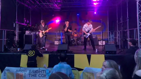 Jon Wright/BBC A music stage with a singer, two guitarists and a drummer. The heads of people watching in the foreground can be seen