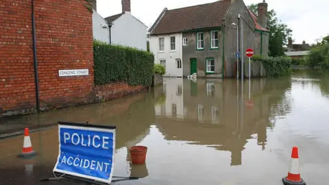 A flooded residential street where muddy water has covered the road and reach the walls of nearby houses. A brick building and hedgerow line the left side, while several older houses with white and green exteriors stand partially submerged. A blue roadside sign reading “POLICE ACCIDENT” is positioned in the foreground, along with traffic cones blocking entry to the flooded area.
