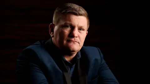 PA Media Ricky Hatton, vestida com uma jaqueta marinha e camisa. Ele tem cabelos escuros curtos e está olhando para a câmera.