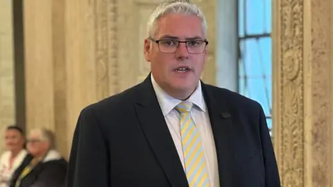 PA Media Gavin Robinson in a black suit and yellow and blue striped tie.
