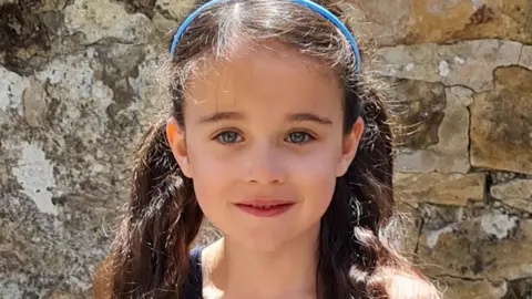 Family handout Head and shoulders of a smiling Millie Gribble in front of a stone wall. She has long dark hair tied back in two bunches with a thin blue headband. 