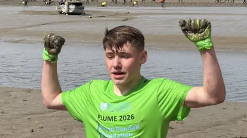 BBC A teenage boy raises his arms in victory as he crosses the finish line of a mud race on a sunny day. He wears a bright green top with matching green gloves with mud on them. His brown hair is wet. People can be seen running through the mud behind him in the distance.