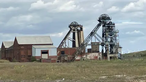 Headstocks at the top of a mine shaft. Next to them is a series of red brick buildings. 