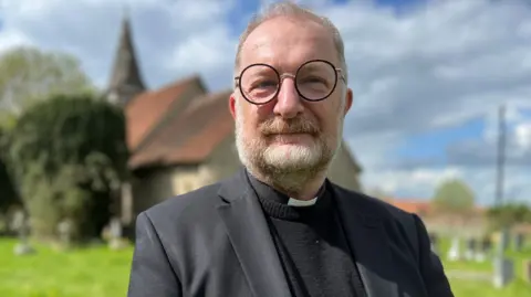 Stuart Woodward/BBC The Reverend Tim Goodbody, a man with short fair hair and a white and ginger beard, wears a black suit with a dog collar. His glasses are black framed and round. A church is out of focus behind him.