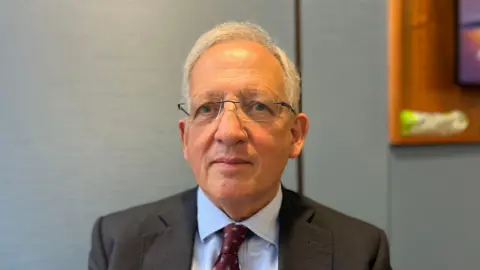 BBC Jon Cunliffe is a white man with grey hair and glasses. He is staring into the camera. He is wearing a dark grey suit with a pale blue shirt and burgundy tie. He is in focus with a blurred blue background behind him.