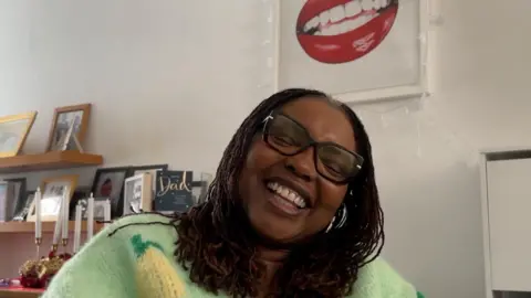 Yvonne Johnson laughing sat in her living room in her wheelchair, with a portrait of her lips behind her.