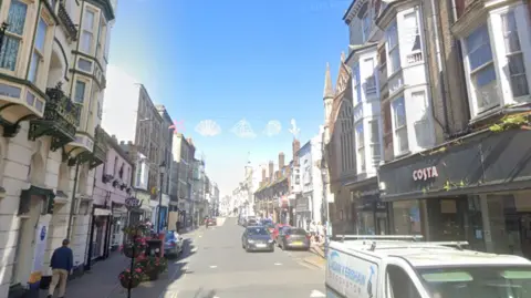 Google A bright, sunlit high street stretches into the distance, lined with rows of older multi-storey buildings housing shops and cafes, including a Costa storefront on the right. Cars are parked along both sides of the road while a few pedestrians walk on the pavements, with decorations strung across the street overhead against a clear blue sky.