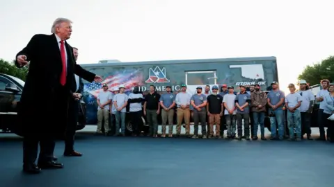 Reuters Trump speaking in front of a line of workmen and a work truck