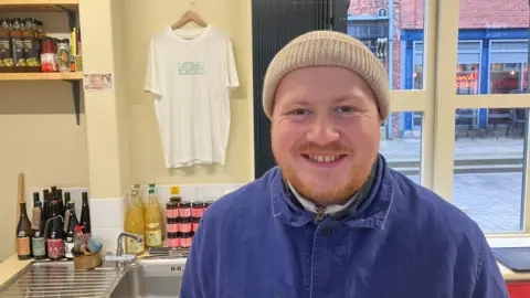 Photograph of Harrison Marslend, who is a trader at Stockport Indoor Market Hall. The 30 year-old is pictured in front of his stall. He is wearing a blue demin overall and a woolly hat. 