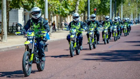 Metropolitan Police A line of Metropolitan Police officers riding high-powered e-bikes down a paved London road in single file formation, wearing full protective gear and high-visibility jackets.