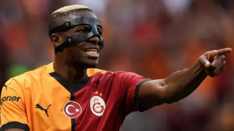 Victor Osimhen, wearing a face mask and a yellow and red Galatasaray shirt, points with his left index finger and his arm extended