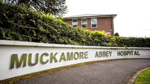 A small white wall has the words 'Muckamore abbey hospital' on it. It sits below a green bush, which runs the length of the wall. Behind the wall is a tall red brick building with three windows on its top floor. A tarmac footpath is in front of the wall. 