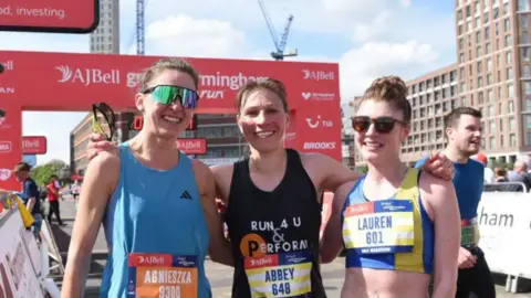 Three women known as Abbey Van Dijk, Agnieszka Wilk and Lauren Cooper smiling at a big red finish line.