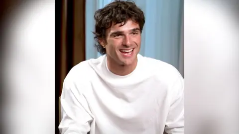 A man, Jacob Elordi, has dark brown hair and wears a white top. He is smiling