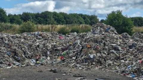 Mounds of rubbish, which looks like a mixture of shredded plastic and dirt, on the floor. Behind it are hedges and trees