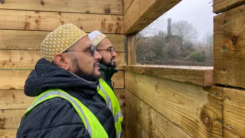 Two men in decorative hats and fluorescent yellow hi-vis waistcoats looking through wooden observation slits