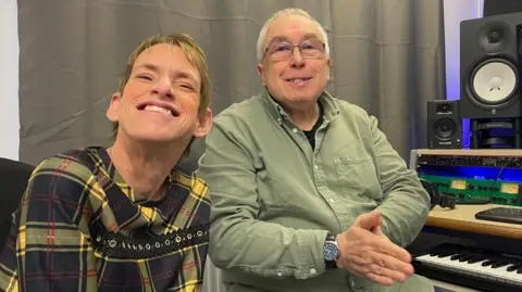 Simon Thake A woman in a yellow dress sits smiling next to a man in a green shirt who sits next to a keyboard