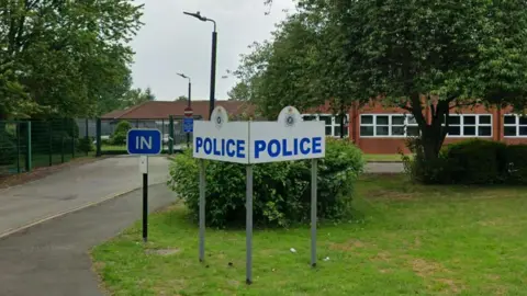 A picture of a police station. There is a white sign in the grounds of the station, that has blue letters spelling police. Behind is a large green tree and behind that is the station.