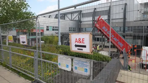 A metal hoarding with a temporary sign pointing to A&E at the Prince Charles Hospital. Through the metal railings a red tipper truck can be seen in front of the main hospital building.