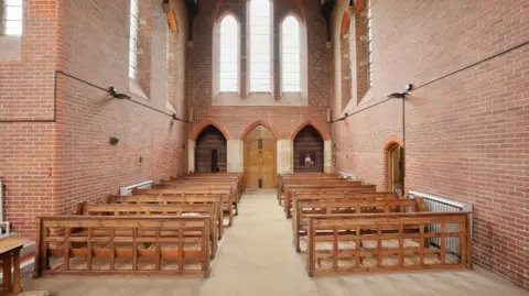 Hunters Estate Agents Rows of wooden benches sit either side of a chapel with an aisle down the middle. At the end is a large wooden door underneath three large-pained glass window frames