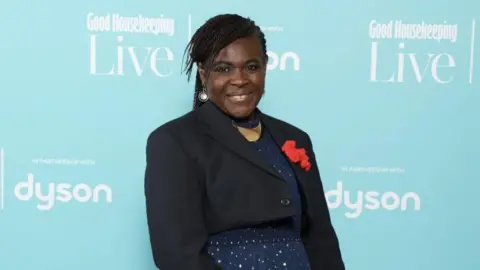 A woman with a pulled-back braided hairstyle, wearing a black jacket and glittery blue dress with a red knitted broach on it. She stands in front of a light blue advertising board at a red carpet event. She is smiling.