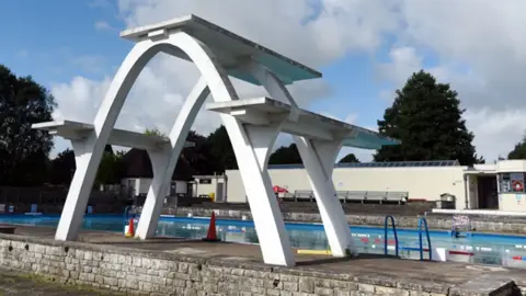 large 1930s white diving board with three boards in front of the outdoor pool in Stroud. Pool has blue steps and lane floats. 