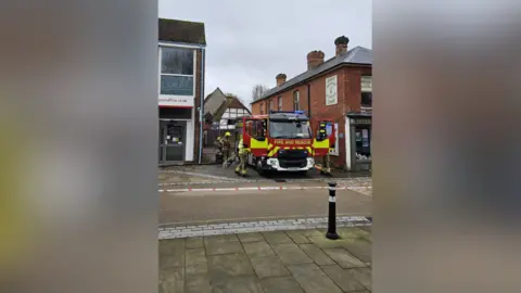 Test Valley Borough Council A fire engine is seen blocking a narrow street in between two small commercial buildings. Fire crews are seen standing around it and red and white tape is seen blocking the street in front. 