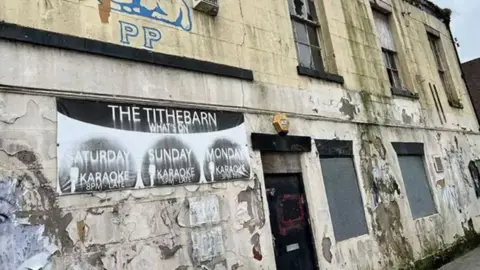 The outside of the Tithebarn pub, a dirty, tired-looking building with peeling white paint, rain damage, boarded up windows and peeling posters